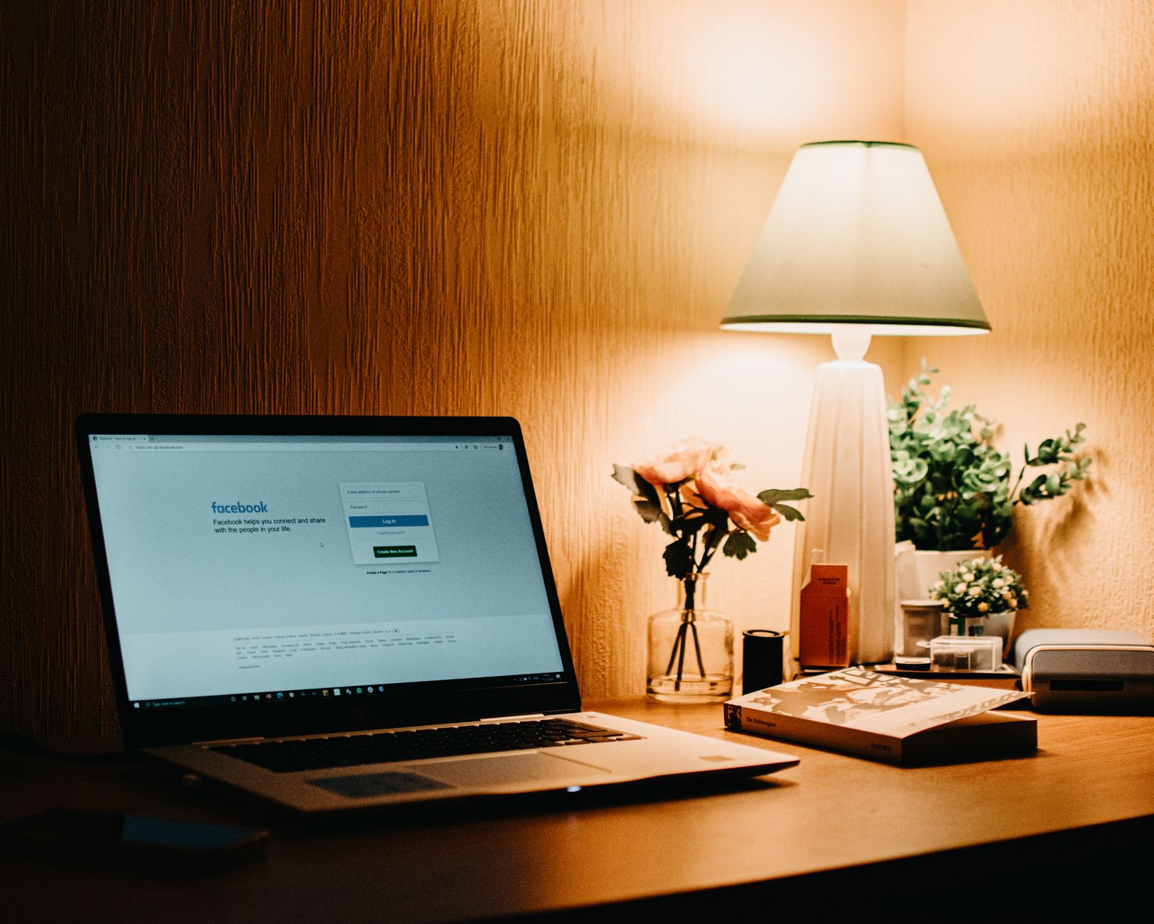 Un bureau chaleureux le soir&nbsp;: un ordinateur portable ouvert, une lampe allumée, un vase de fleurs et un livre posé sur la table.