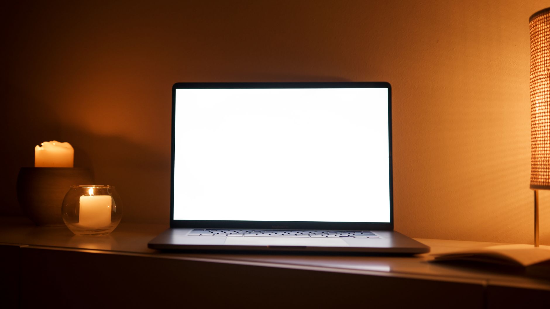 Un poste de travail à la maison, le soir&nbsp;: un ordinateur portable ouvert entre des bougies allumées et une lampe.